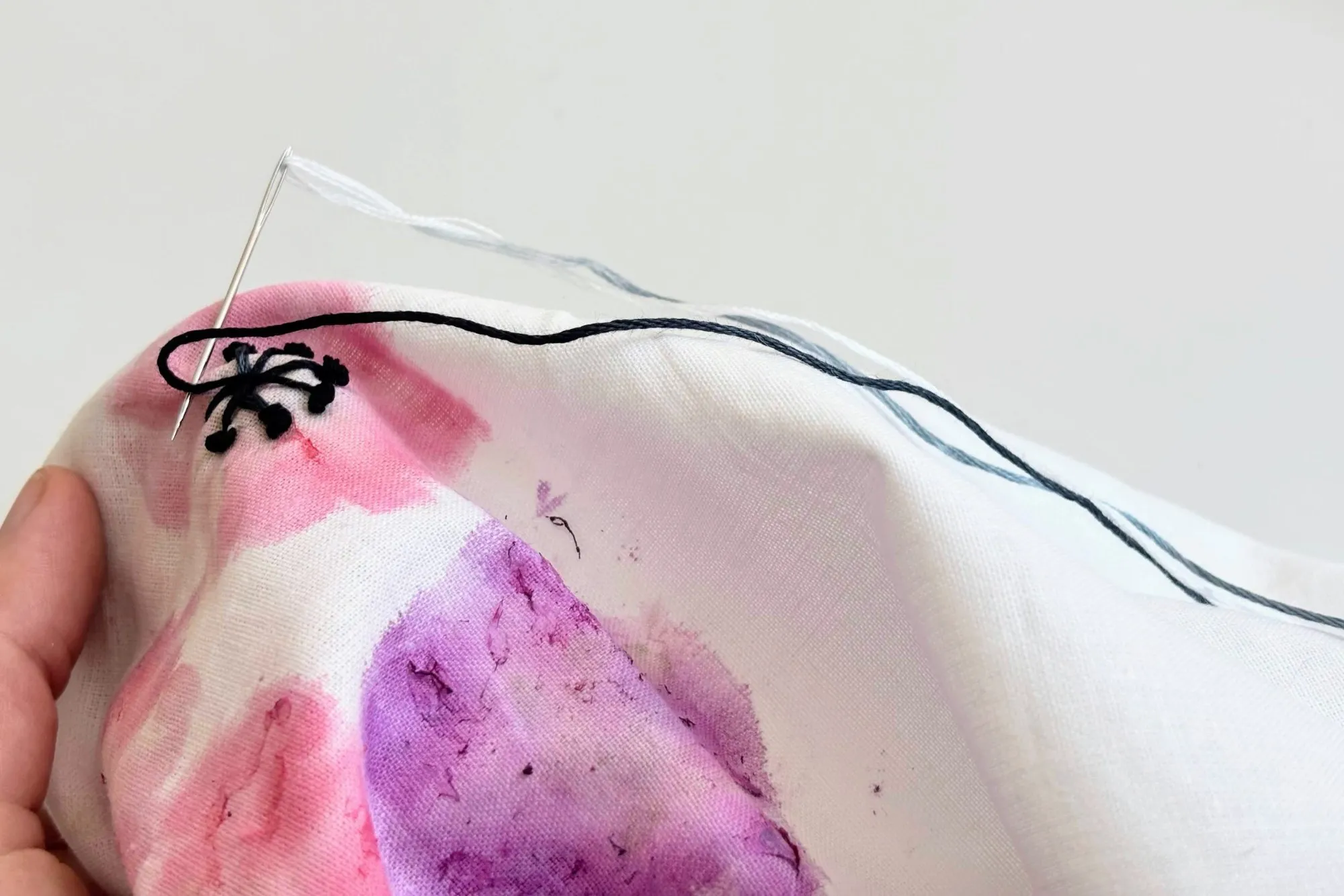 Hands holding hand-dyed fabric with free-motion embroidery in progress — needle, thread and a small stitched motif, by Zane Shumeiko